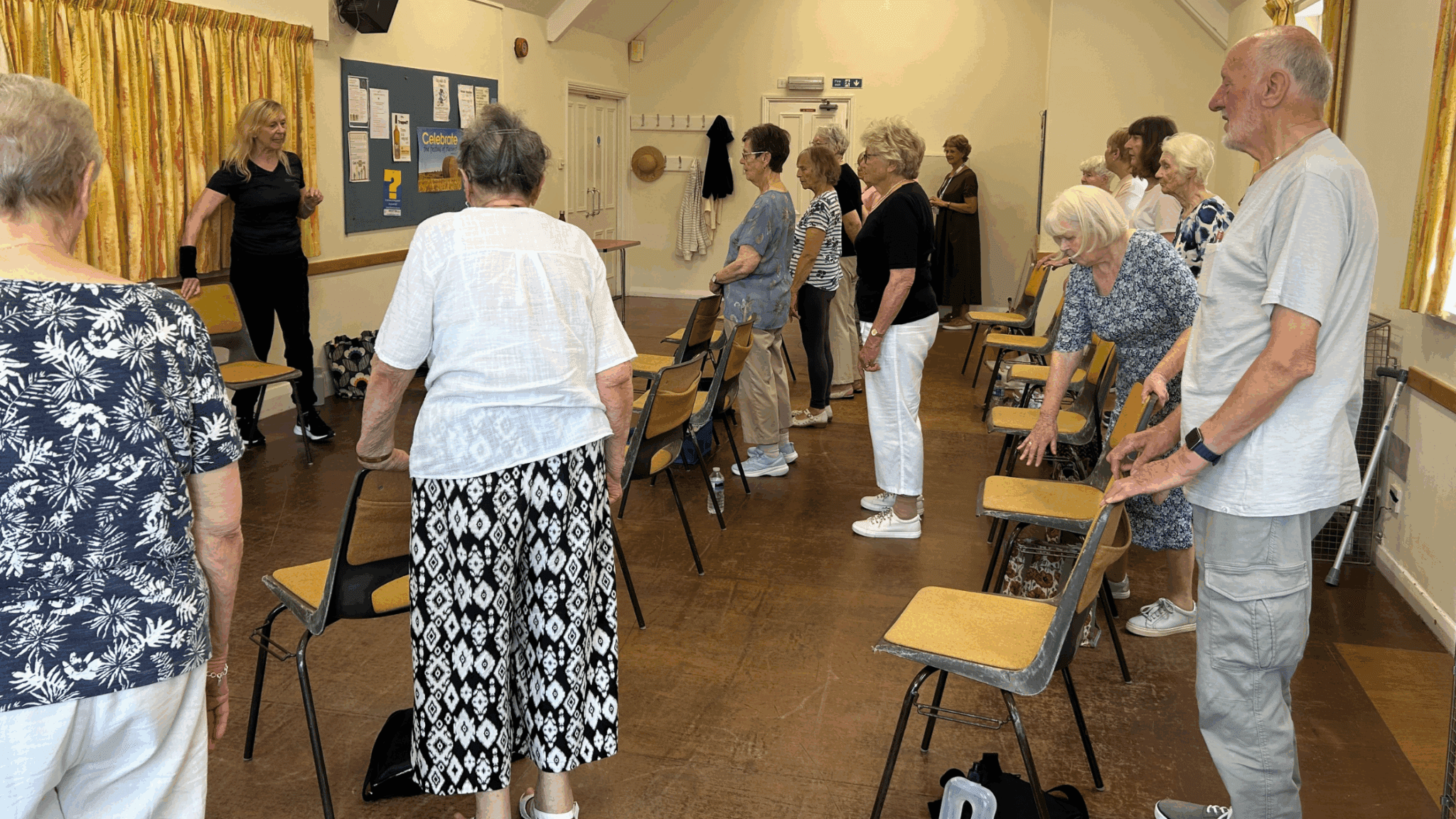 A group of seniors in a room standing with chairs as they participate in an exercise class